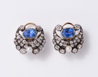 May include: A pair of earrings featuring a central oval blue sapphire surrounded by numerous small clear stones set in a dark metal frame. Each earring includes a single white pearl at the bottom and a curved gold-toned wire hook at the top.