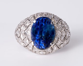 May include: A silver-toned ring featuring a large, oval-cut, deep blue gemstone at the center. The band is wide and ornate, covered in a dense, intricate pattern of small, clear, sparkling stones set within a metallic, filigree-style framework.