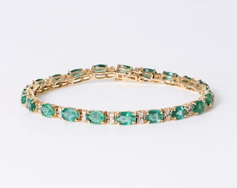 May include: A gold tennis bracelet featuring a series of oval-cut green emeralds alternating with small, clear round-cut diamonds. The gemstones are set in a yellow gold metal frame with a secure clasp mechanism visible at the top center.