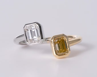 May include: A bypass ring featuring two emerald-cut stones in bezel settings. One stone is clear and set in a silver-toned band, while the other is a deep yellow stone set in a gold-toned band. The two bands overlap to create a continuous design.