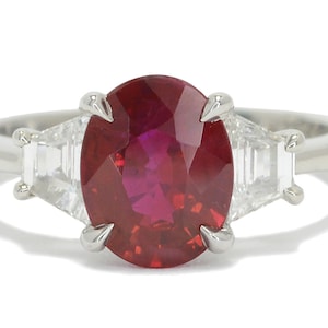 May include: A platinum ring with a large oval ruby gemstone and two smaller baguette-cut diamonds on either side.