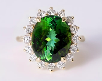 May include: A yellow gold ring featuring a large, oval-cut, deep green gemstone at the center. The central stone is surrounded by a halo of sixteen small, round, clear white diamonds set in a floral-like arrangement with four gold prongs.