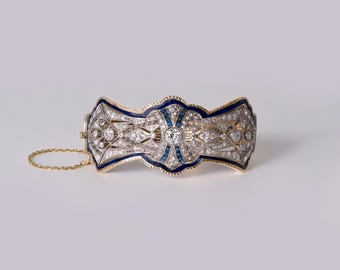 May include: A gold hinged bracelet featuring an intricate openwork design set with numerous small diamonds and deep blue enamel accents. The piece includes a central round diamond, baguette-cut blue stones, and a delicate gold safety chain attached.