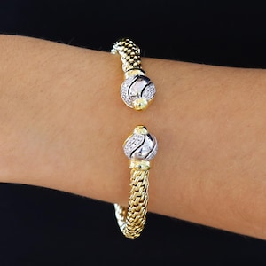 May include: A gold-toned bracelet with a woven chain design. The bracelet features two decorative ends with a silver and diamond-like pattern, and gold accents. The bracelet is open, allowing it to be easily put on and taken off.