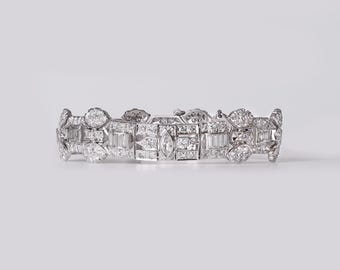 May include: A silver-toned metal bracelet featuring a repeating geometric pattern set with numerous small, clear, faceted stones. The design includes rectangular baguette-cut stones and a central marquise-cut stone, all arranged in a symmetrical link structure.