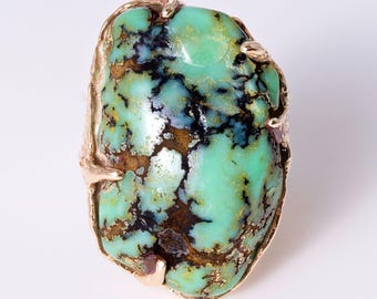 May include: A ring featuring a large, irregular, polished turquoise stone with prominent black and brown matrix veining, set in a textured, organic-shaped gold-toned metal prong setting that follows the natural contours of the gemstone.