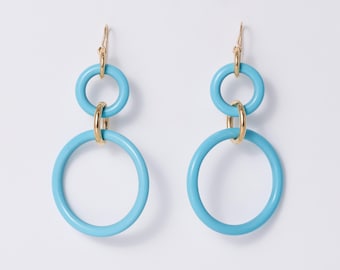 May include: A pair of drop earrings featuring two light blue circular rings of different sizes connected by a small gold-toned metal ring. The earrings hang from gold-toned metal hooks, creating a layered, geometric design with a smooth finish.
