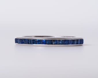 May include: A silver-toned metal band ring featuring a continuous channel setting of small, square-cut deep blue sapphires that wrap around the entire circumference of the band, creating a uniform and polished appearance with a smooth metallic finish.