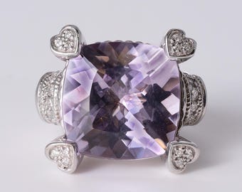 May include: A silver ring featuring a large, cushion-cut, light purple amethyst gemstone at the center. The setting includes four heart-shaped accents at the corners, each pavé-set with small clear stones, and a band decorated with clear stones.