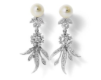 May include: A pair of silver-toned drop earrings featuring a round white pearl at the top, followed by a small cluster of clear stones, and a dangling floral-inspired element adorned with multiple small clear stones set in a metallic frame.