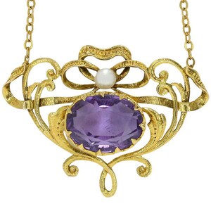 Könnte beinhalten: Ein goldener Anhänger mit einem oval geschliffenen violetten Amethysten in einem verzierten, texturierten Goldrahmen mit geschwungenen Metallelementen und einer einzelnen weißen Perle als Akzent an der Oberseite des Designs.