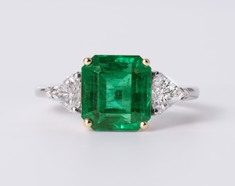 May include: A ring featuring a large, rectangular emerald-cut green gemstone set in gold prongs, flanked by two triangular-cut clear diamonds on each side, all mounted on a polished silver-toned metal band against a plain white background.