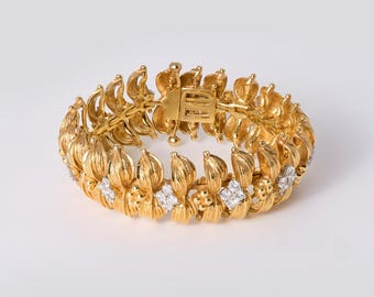 May include: A gold-toned bracelet featuring a repeating leaf-like pattern with textured surfaces. The design includes small, clear, square-cut stones embedded at intervals throughout the circular band, which is secured by a rectangular metal clasp.
