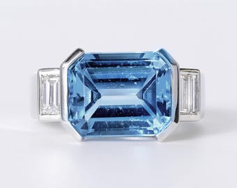 May include: A silver-toned ring featuring a large, rectangular, emerald-cut light blue gemstone set in the center. The central stone is flanked on each side by a smaller, rectangular, clear baguette-cut gemstone set into the metal band.