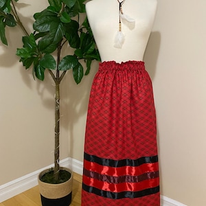 May include: A red plaid skirt with black and red ribbon detailing. The skirt features a gathered waistband and is displayed on a mannequin. A brown necklace with feathers is also visible. A potted plant is in the background.