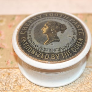 May include: A vintage white ceramic jar of Cherry Tooth Paste. The lid features a circular label with text and a profile of a woman. The text reads "Patronized by the Queen" and "Prepared by John Gosnell & Co London".