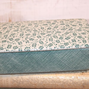 May include: A rectangular ceramic box with a lid, featuring a teal and white color scheme. The lid is adorned with a delicate green leaf pattern, while the box's base has a textured teal design. The box is resting on top of a stack of vintage books.