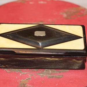 May include: A rectangular box with a dark brown base and a lid featuring a cream-colored border surrounding a dark brown diamond shape. A small, rectangular silver-toned metal inlay is centered within the dark brown diamond on the lid.