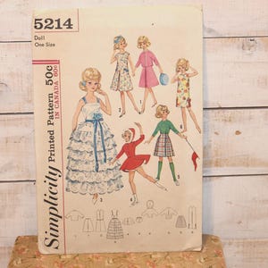 May include: Vintage Simplicity pattern 5214 for a doll, featuring illustrations of the doll in various outfits. The pattern cover displays the text "Doll One Size" and "Printed Pattern 50c in Canada 60c". The background is a light wood.