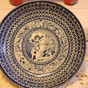 May include: A blue and white ceramic saucer with a floral design and a central image of a figure holding a flower. The saucer has a repeating pattern of small blue dots around the edge.