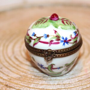 May include: A small, round, white porcelain trinket box shaped like a fruit, featuring hand-painted green leaves, red accents, and small blue flowers. The box has a brass-coloured metal hinge and a small red knob on top for opening the lid.