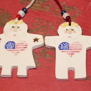 May include: Two white ceramic ornaments shaped like figures with outstretched arms. Each figure features a painted heart with red stripes and a blue field with white dots. They have yellow hair, green eyes, and are suspended by strings with red, white, and blue beads.