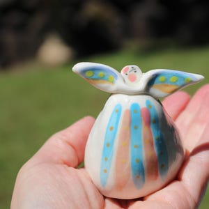 May include: A small white ceramic figurine shaped like an angel with outstretched wings. The body features vertical stripes in light blue and peach, accented with small yellow dots. The wings have blue edges and yellow spots, with a pink face.
