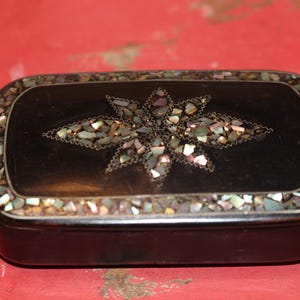 May include: A rectangular black box featuring an inlaid star-shaped design on the lid, composed of small, iridescent, multi-colored shell fragments. The perimeter of the lid is also decorated with a border of similar shell pieces.