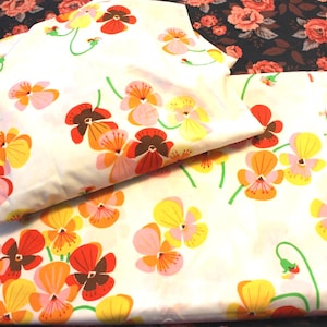 May include: A folded white fabric featuring a repeating pattern of stylized pansy flowers in shades of orange, yellow, pink, and dark brown with green stems. The fabric is layered, showing a consistent floral print across the visible surfaces.