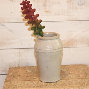 May include: A light gray ceramic vase with a cylindrical shape and a wide rim. The vase holds a stem of faux succulents in shades of red and green. The vase is placed on a floral patterned surface against a white wooden background.