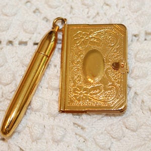May include: A gold-toned metal charm set featuring a miniature book with an embossed floral pattern and a smooth, rounded pen-shaped charm. Both items are connected by a small circular jump ring, creating a cohesive, polished metallic accessory.