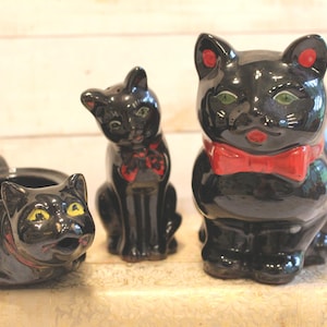 May include: A set of three black ceramic cat figurines. The set includes a creamer, a salt shaker, and a sugar bowl. The cat figurines have red bow ties and accents, with yellow eyes and red accents on the ears.