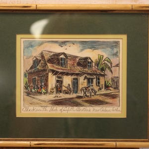 May include: A framed print featuring a colored sketch of a building labeled Blacksmith Shop of Lafitte Brothers New Orleans, La. The artwork is encased in a dark green mat and a light-colored bamboo-style frame with a rectangular profile.