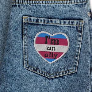May include: Blue denim jeans with a heart-shaped patch that says "I'm an ally" in black text. The patch has a pink, blue, and white color scheme.