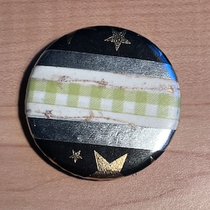 Pode incluir: Botão redondo com fundo preto e detalhes de estrelas douradas. O botão apresenta listras horizontais prateadas, brancas e um padrão xadrez vichy verde claro. Acessório decorativo.