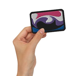 Pode incluir: Patch bordado retangular com borda preta. O patch apresenta um design abstrato com padrões em redemoinho em branco, rosa, roxo e azul sobre um fundo preto. O patch tem cantos arredondados.