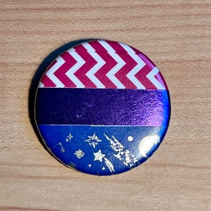 May include: A round button with a pink and white chevron pattern, a purple stripe, and a blue background with gold glitter stars and a crescent moon.