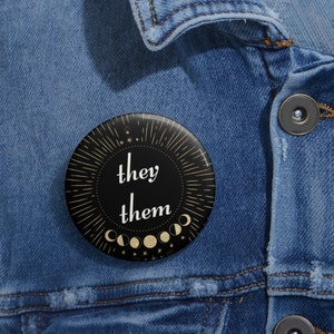 May include: A circular black badge with the words "they them" in white script. The badge has a gold sunburst design, moon phases, and stars. It is pinned to a denim jacket.