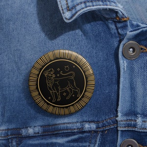 Puede incluir: Un botón negro y dorado con un dibujo de línea de un toro, el símbolo astrológico de Tauro, rodeado de estrellas. El botón está en una chaqueta vaquera.