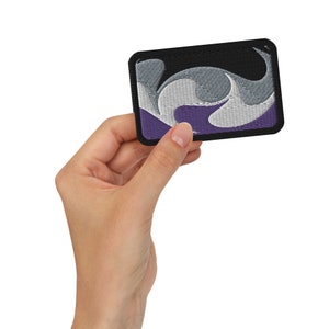May include: An embroidered patch with a black border, featuring a stylized wave design in purple, grey and white.