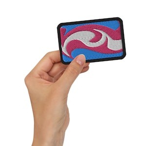 Puede incluir: Un parche bordado azul, blanco y rosa con un diseño de espiral. El parche tiene un borde negro y se sostiene en una mano.