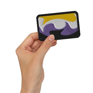 May include: A purple, white and yellow embroidered patch with a black border. The patch is being held by a hand.