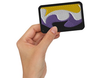 May include: A purple, white, and yellow embroidered patch with a black border. The patch is being held by a hand.
