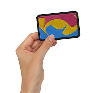 May include: A small embroidered patch with a blue, yellow and pink swirl design. The patch has a black border and is held in a hand.