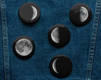 May include: Five circular pin-back buttons featuring high-contrast lunar phase illustrations. Each button displays a different moon phase, including a full moon, a crescent moon, and quarter moons, rendered in shades of gray against a black background.