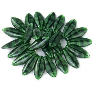 May include: A collection of dark green, elongated beads with a striped pattern, arranged in a circular shape against a white background. These beads could be used for jewelry making or other craft projects.