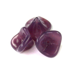 Puede incluir: Tres cuentas de vidrio de color púrpura con forma de flor. Las cuentas son translúcidas y tienen un acabado brillante.