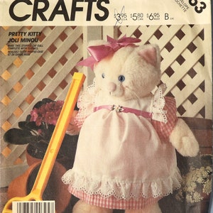 May include: A vintage McCall's Crafts pattern envelope featuring a stuffed cat doll named "Pretty Kitty Joli Minou." The doll wears a pink dress with lace trim and a matching bow. The pattern includes instructions to make the doll with clothes.