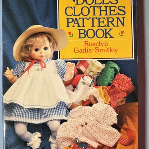 May include: A book titled Dolls' Clothes Pattern Book by Roselyn Gadia-Smitley. The cover features a doll wearing a blue and white gingham dress, a white pinafore, and a straw hat, alongside fabric scraps, a wooden spool, and a red pincushion.