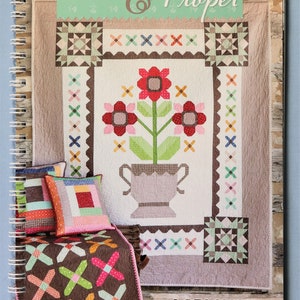 May include: A spiral-bound instructional book titled Prim & Proper by Lori Holt of Bee in My Bonnet Co. The cover features a quilt pattern with a central flower vase design, surrounded by smaller decorative pillows and a brown patterned quilt.